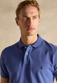 Zils polo krekls, kas izgatavots no teksturēta auduma, ar klasisku apkakli, divām pogām un mazu tumšu logotipu kreisajā krūtī.