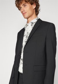 Schwarze Anzugjacke mit Revers und zwei Knöpfen, kombiniert mit einem weißen Hemd im schwarzen Marmor-Muster. Glattes Gewebe, taillierte Passform.