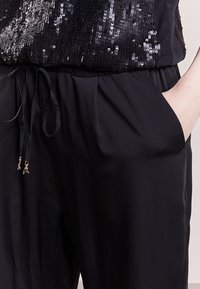 Mono negro con parte superior de lentejuelas, que cuenta con un cinturón ajustable. Tejido suave, bolsillos laterales y acentos metálicos en el cordón.