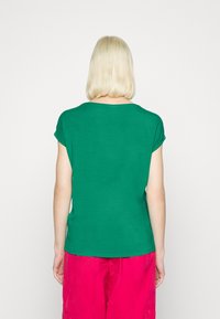 Zelená košile s krátkým rukávem a volnějším střihem, kulatým výstřihem a hladkou texturou, v kombinaci s jasně růžovými volnými kalhotami.