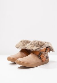 Paire de bottines beige avec des bordures en fausse fourrure, des lanières en cuir et des semelles en caoutchouc, disposées côte à côte sur une surface blanche.