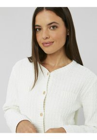 Cardigan blanc en tricot avec col et poignets côtelés, doté d'une fermeture à boutons et d'un tissu texturé. Petite poche passepoilée sur le côté gauche.