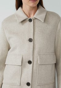 Beige wollen blend jas met een kraag, vier zwarte knopen en twee voorzakken. De stof heeft een zachte, gestructureerde uitstraling.