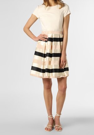 Femme portant une robe à manches courtes, longueur genoux, avec un haut beige et une jupe plissée rayée crème, rose et noire, assortie à des sandales à talons beiges.