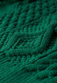 Tissu tricoté vert présentant des motifs en relief et des tresses. Le fil semble doux et épais, avec un aspect cosy et superposé.