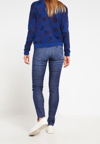 Mujer vestida con un suéter de patrón floral azul, jeans oscuros ceñidos y zapatillas blancas, de pie con la espalda hacia el espectador.