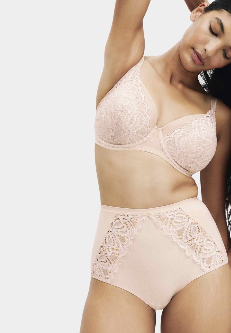 Kvinde iført nøgenfarvet blonde-bh og trusser med høj talje, løfter den ene arm og har lukkede øjne mod en ensfarvet, lyse baggrund.