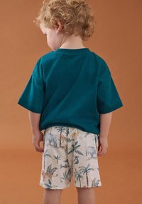 T-shirt à manches courtes couleur sarcelle associé à un short crème orné d'un imprimé d'animaux de la jungle colorés et de motifs de palmiers. Texture lisse partout.