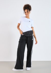 Juodas siuvinėtas fanny pack‘as nešiojamas ant baltos marškinėlių ir juodų sportinių kelnių su baltomis juostelėmis. Modelis stovi neutralioje fone.
