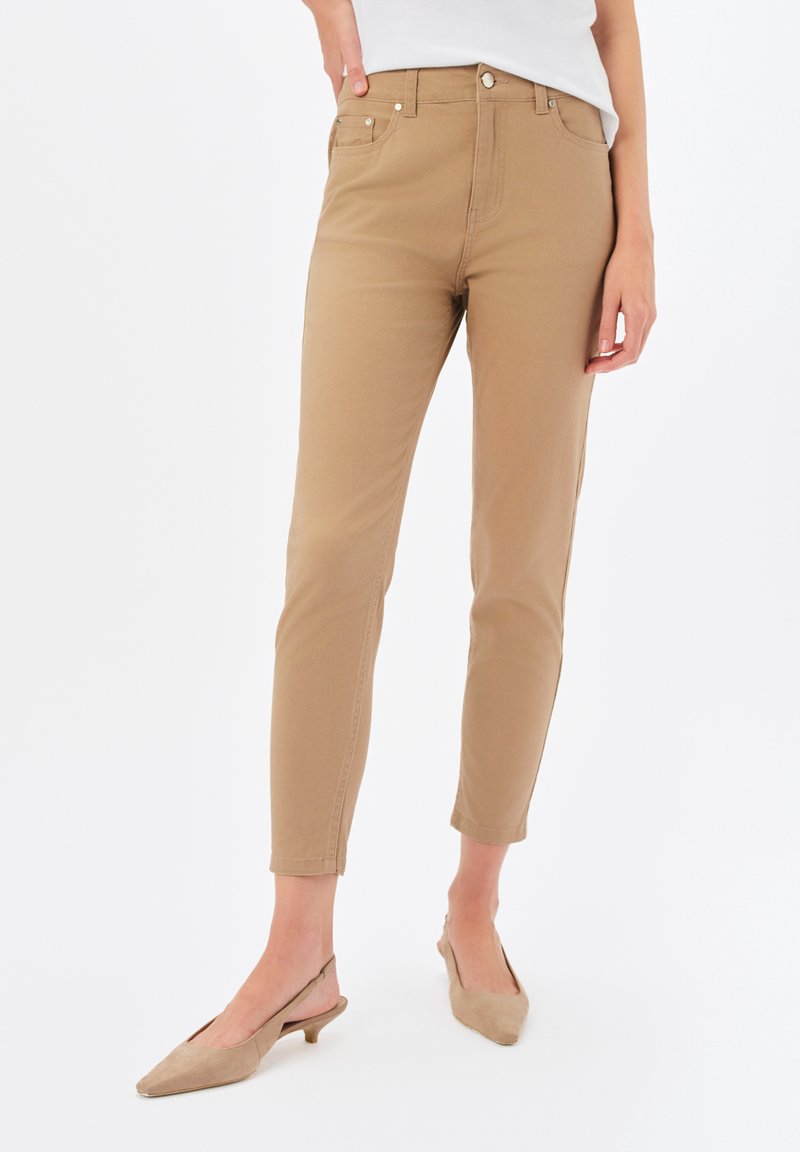 Beżowe spodnie do kostek o dopasowanym kroju, wykonane z mieszanki bawełny, z przednimi kieszeniami i zapięciem na guzik. Noszone z beżowymi butami na obcasie typu slingback.