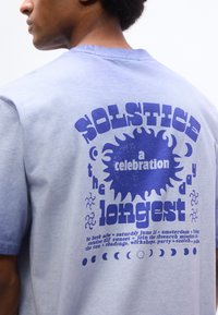 Άνδρας που φοράει γαλάζιο πουκάμισο με σκούρο μπλε κείμενο για τον εορτασμό του ηλιοστασίου, γραφικό ήλιου, φάσεις της σελήνης και λεπτομέρειες εκδήλωσης στο πίσω μέρος.