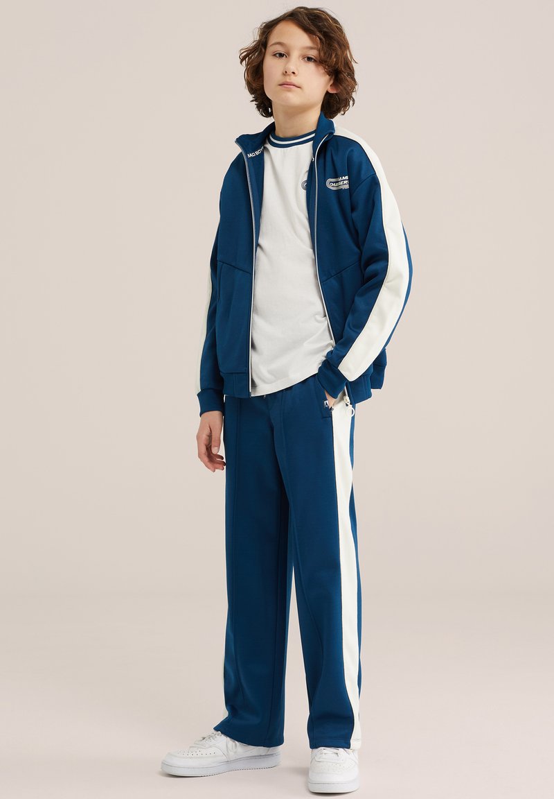 Ung person står iført blå og hvid træningsjakke, matchende bukser, hvid T-shirt og hvide sneakers mod ensfarvet baggrund.