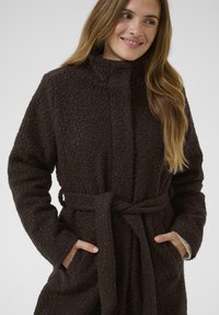 Abrigo de un marrón oscuro, texturizado, con un cuello alto, mangas largas y un lazo en la cintura. Presenta bolsillos laterales y un material suave y peludo.
