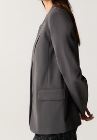 Blazer gris avec une coupe ajustée, texture lisse, revers crantés, poche arrière simple et manches longues, vu de côté.