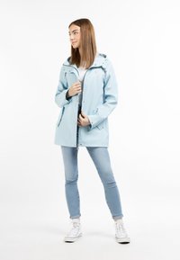 Jachetă impermeabilă de color bleu deschis, cu glugă, închidere cu fermoar și nasturi. Are două buzunare frontale și manșete ajustabile. Purată peste un top alb.