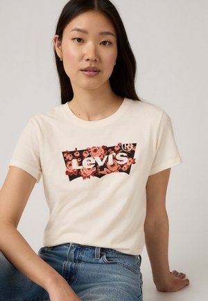 T-shirt en coton crème avec un logo floral Levi's en rouge et noir, à manches courtes, et un design classique à col rond, associé à un jean bleu.
