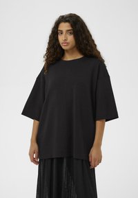 Schwarzes übergroßes T-Shirt mit kurzen, breiten Ärmeln und rundem Ausschnitt, aus glattem Stoff. Kombiniert mit einem langen schwarzen Rock.