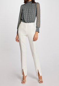 Weiße, maßgeschneiderte Hosen mit Schlitzsäumen, kombiniert mit einer transparenten, gemusterten blauen Bluse mit hohem Kragen und langen, puffärmel.