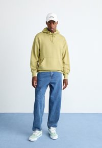 Giovane in felpa verde chiaro, cappellino bianco Dickies, jeans blu e sneakers verde chiaro e bianche, in piedi su un pavimento blu contro una parete bianca.