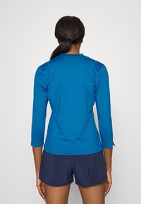 Osoba z ciemnymi włosami związanymi w kucyk, ubrana w niebieską koszulę z długim rękawem i granatowe szorty, stojąca tyłem na jednolitym tle.