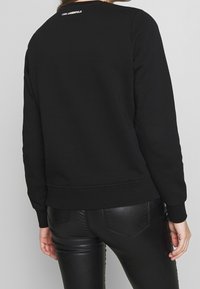 Felpa nera in cotone con collo rotondo, polsini e orlo a costine. Presenta un piccolo logo bianco sulla parte superiore della schiena. Indossata con pantaloni neri lucidi.