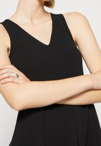 Czarna sukienka bez rękawów z dekoltem w serek, wykonana z gładkiego materiału, z dopasowanym krojem w górnej części i plisowaniem w talii.