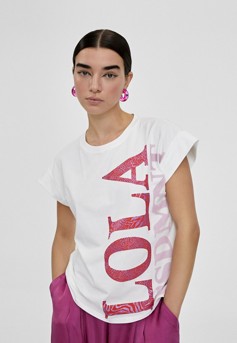 Vit bomulls-T-shirt med oversized passform, med iögonfallande röda och rosa textmönster. Modellen bär rosa cirkulära örhängen, styla med lila byxor.