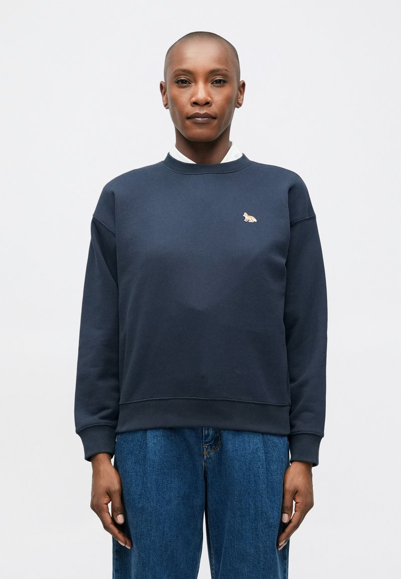 Tumši zils sporta krekls, izgatavots no mīkstas auduma, ar apaļu kakla izgriezumu, ribotām aprocēm un mazu izšuvumu uz krūškurvja.