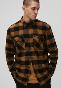 Brązowa i czarna koszula z flaneli w kratę z zapięciem na guziki, dwoma kieszeniami na piersi oraz metką marki na lewej kieszeni. Długie rękawy i luźny krój.