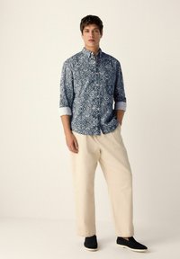 Herren Hemd in Blau mit Muster und hochgerollten Ärmeln, kombiniert mit cremefarbenen Hosen und schwarzen Slippern. Einfaches Design, strukturierter Stoff.