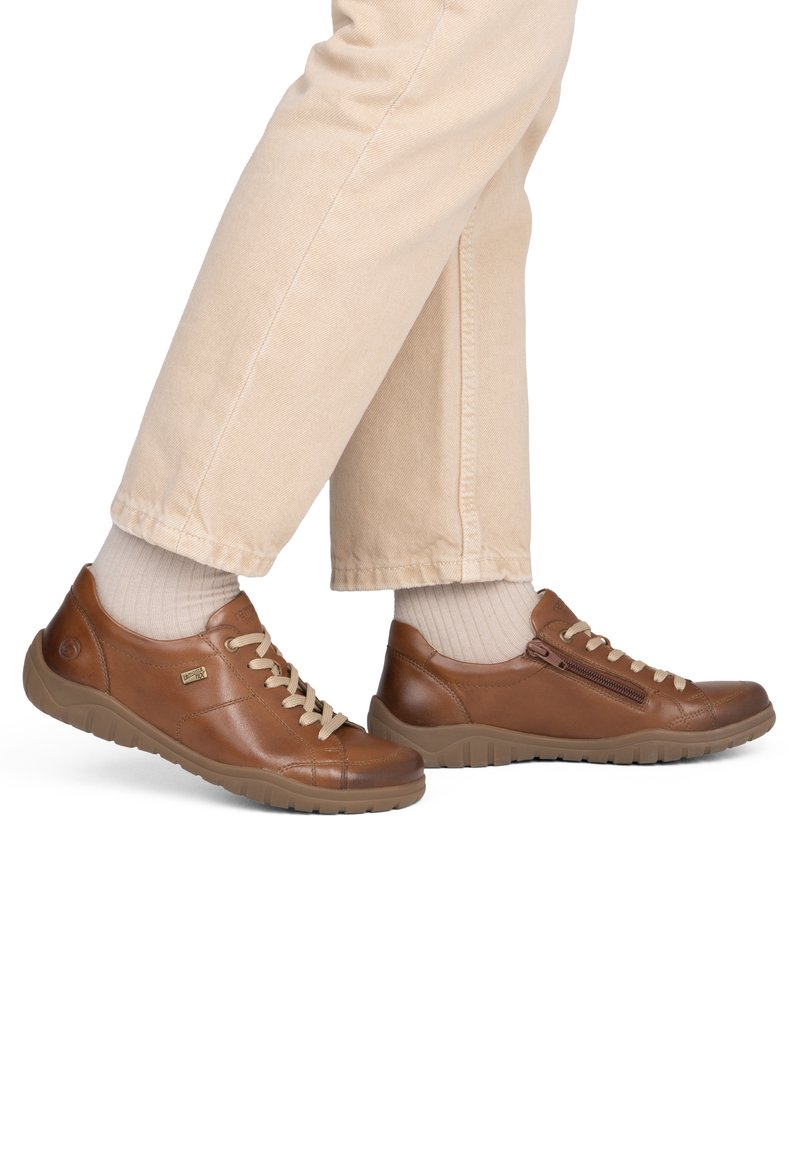 Personne portant un pantalon beige, des chaussettes écrues et des baskets en cuir marron à lacets avec fermetures éclair latérales, debout avec un pied en avant.