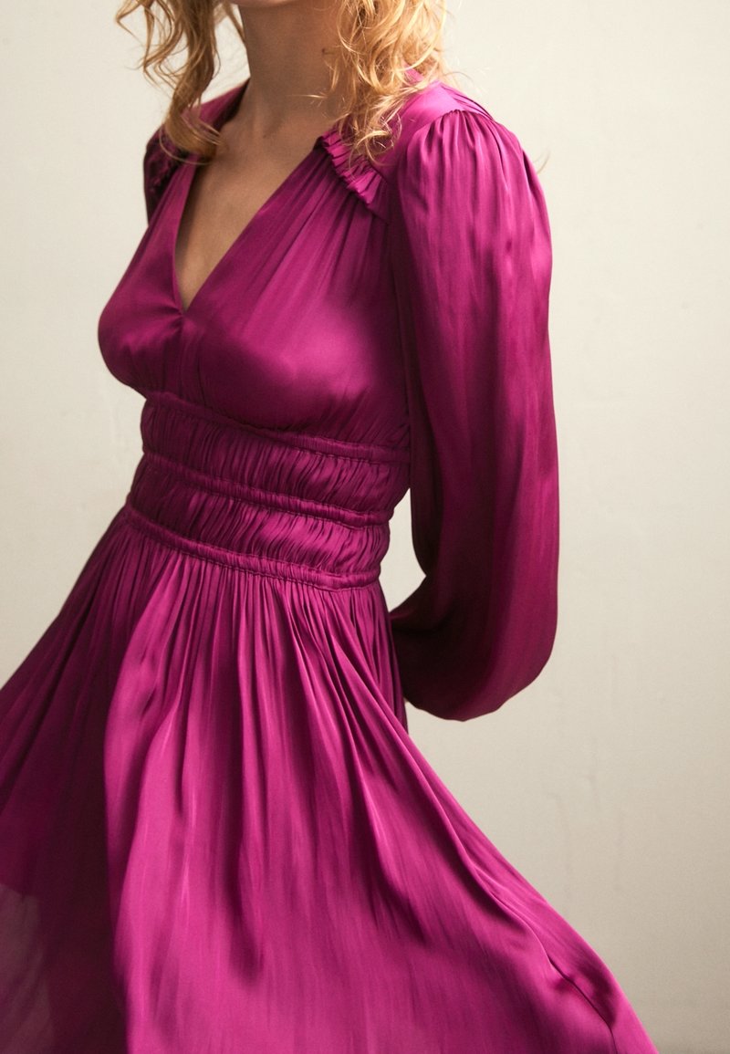 Fuchsia jurk met poflange mouwen, een V-hals en een gerimpelde taille. Gemaakt van lichtgewicht, vloeiende stof met een gladde textuur.