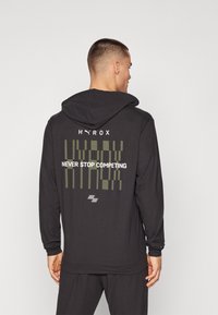 Svart langermet hettegenser med hette, grønt grafisk design på baksiden med teksten "HYROX" og "NEVER STOP COMPETING". Glatt tekstur.
