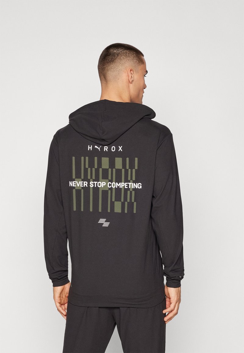 Svart langermet hettegenser med hette, grønt grafisk design på baksiden med teksten "HYROX" og "NEVER STOP COMPETING". Glatt tekstur.