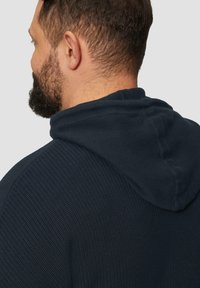 Marineblaue Strickkapuzenjacke mit einer strukturierten Oberfläche, ausgestattet mit einer großen Kapuze und sichtbaren Nähten an den Schultern und dem Rücken.