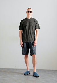 Mørkegrøn t-shirt, sort og hvid stribede shorts og blå slip-on sko. Modellen har solbriller på og står på en grå overflade med en ensfarvet baggrund.