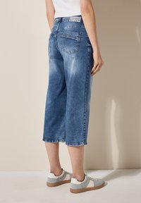 Denimové culoty v středně modré barvě, se uvolněným střihem, zkrácenou délkou, jemným vyblednutím a zadními kapsami. Spárované se šedými a bílými botami.