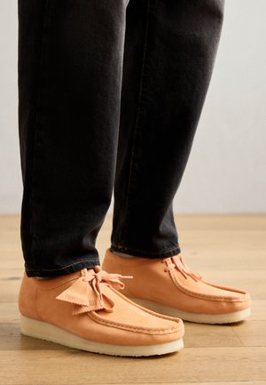 Tan suede moccasin shoes with a white crepe sole, featuring stitched detailing and bow accents, paired with dark jeans.