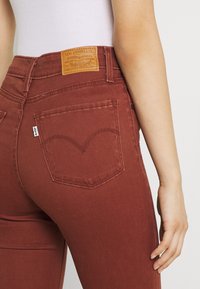 Jeans de mezclilla color óxido con una cintura alta, que presentan un parche de cuero con el logo y un característico cosido curvado en los bolsillos traseros.