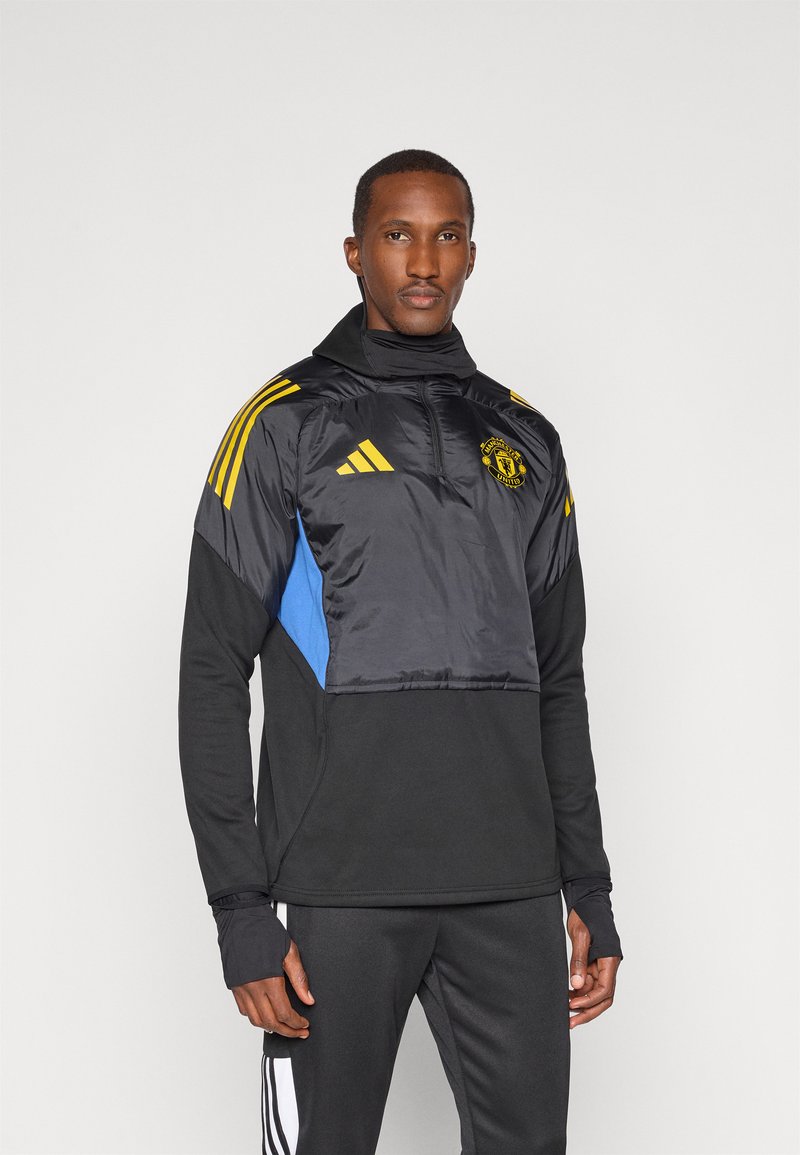 Schwarze und blaue Pulloverjacke mit hohem Kragen, ausgestattet mit gelben Adidas-Streifen und einem Manchester United-Logo auf der Brust. Glattes Material.