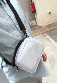 Sac bandoulière blanc et glossy de forme rectangulaire, avec une bandoulière noire et un discret logo sur le devant. Porté sur une chemise bleue texturée.