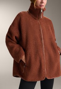 Brown fleece jacket with high collar, full zipper, side pockets, and smooth leather accents along the zipper and hem. Soft, textured fabric.