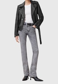 Vrouw in een zwarte leren motorjack, witte t-shirt, hooggeplaatste grijze spijkerbroek met zwarte riem, en zwarte puntige kitten heels.