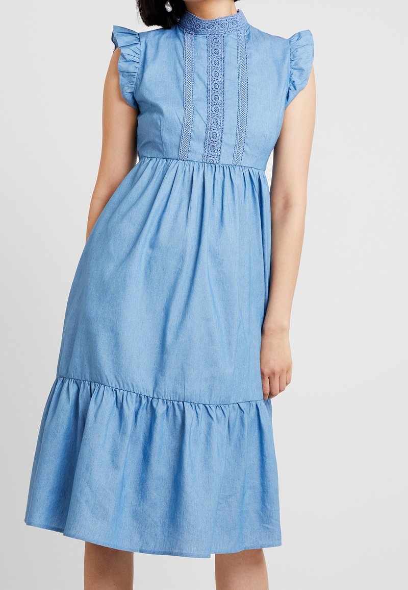 Světle modrá džínová šaty s vysokým límcem, krátkými volánovými rukávy, vyšívanými detaily a rozšířeným volánkovým lemem. Hladká textura.
