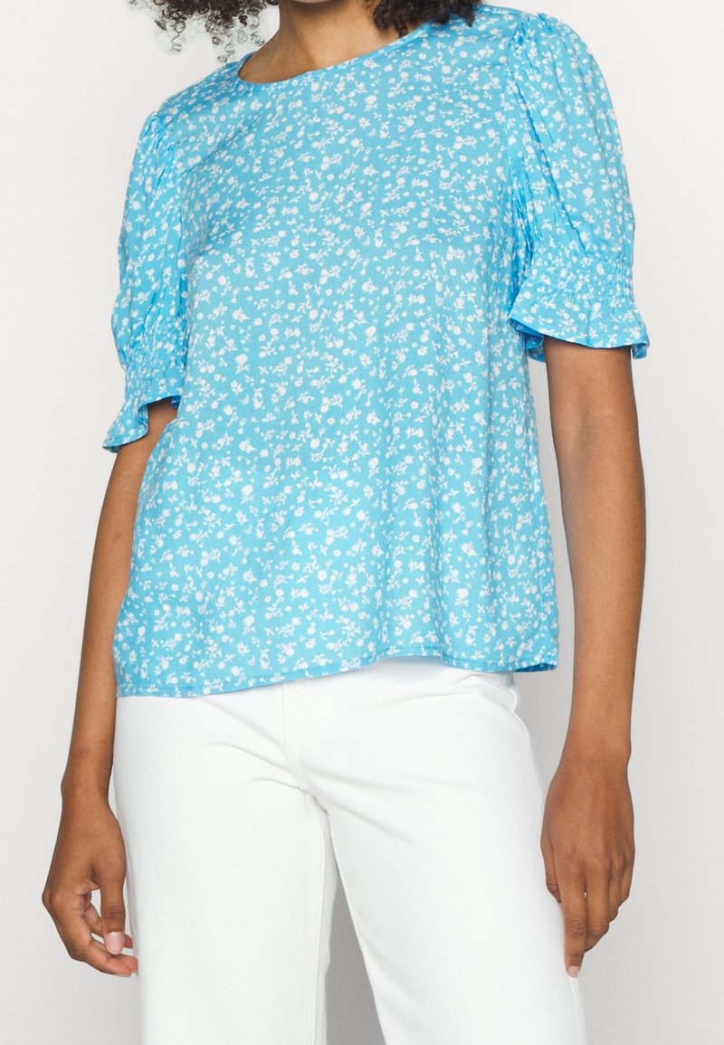 Blusa azzurro chiaro con motivo floreale, maniche corte a palloncino, scollo rotondo e vestibilità rilassata, abbinata a pantaloni bianchi.