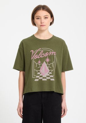 Jeune femme portant un t-shirt Volcom vert olive avec un motif géométrique rose et des étoiles, associée à un pantalon noir, se tenant devant un fond blanc.