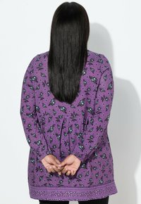 Fioletowa tunika w kwiaty z długimi rękawami, posiadająca marszczoną linię talii, kontrastujące czarne kwiaty oraz ozdobny brzeg. Pokazany widok od tyłu.