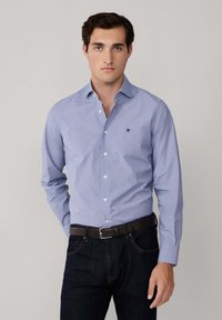 Uomo che indossa una camicia azzurro chiaro con bottoni, jeans scuri e una cintura nera, in piedi davanti a uno sfondo chiaro e uniforme.