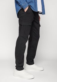 Zwarte cargobroek met zijzakken, gemaakt van stevig materiaal. Gecombineerd met witte sneakers en een denim overhemd met knoopsluiting.