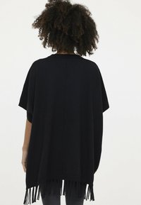 Schwarzer Poncho mit kurzen Ärmeln, lockerem Schnitt und Fransen entlang des Saums. Hergestellt aus einem weichen, strukturierten Stoff.